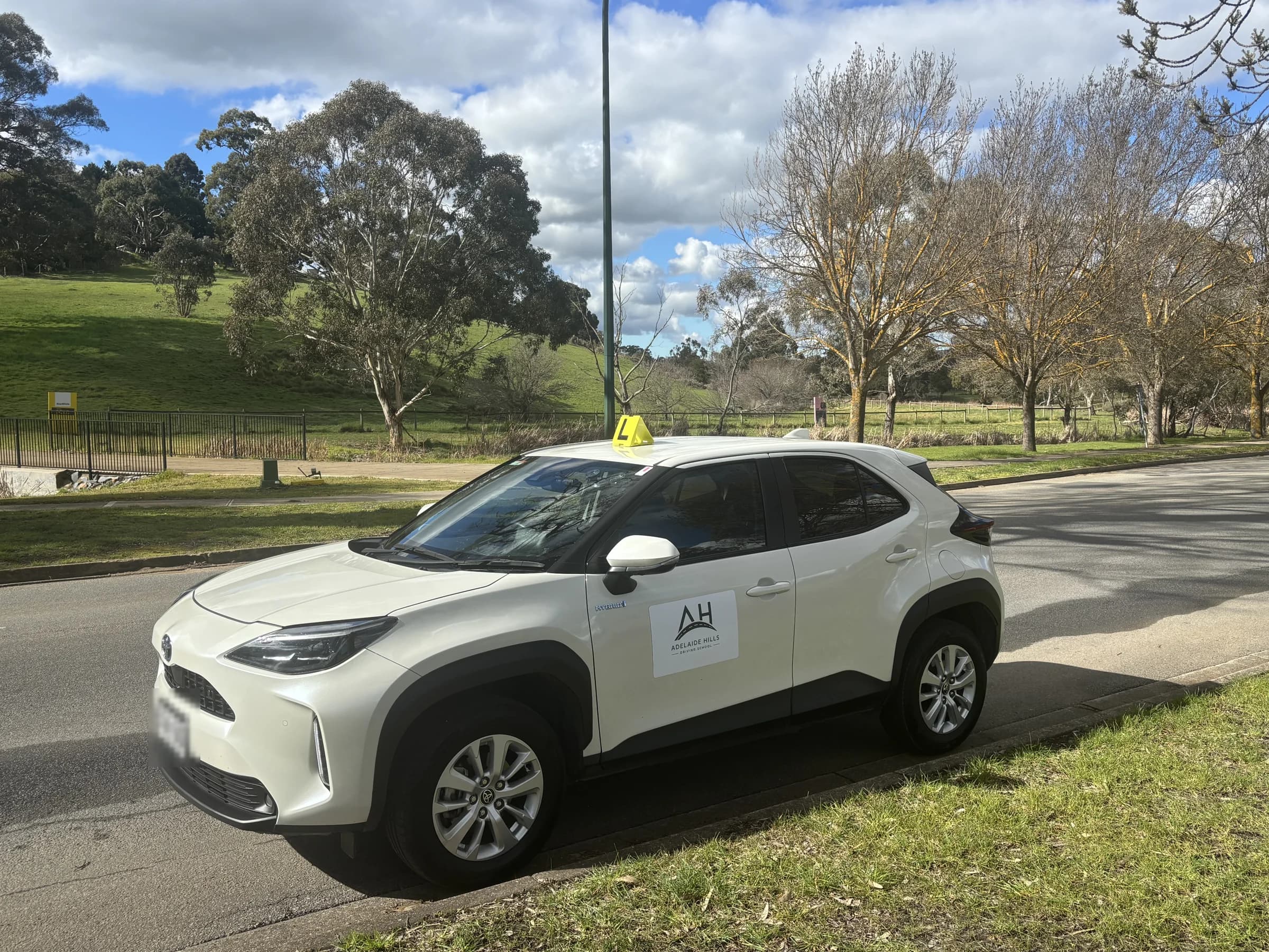 The Adelaide Hills Driving School dual-control Toyota Yaris Cross, signed with the AH logo, parked among gum trees on a quiet sunlit road